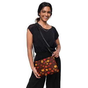 May include: A brown crossbody bag with a colorful autumn leaf pattern. The bag has a black adjustable strap and a flap closure.