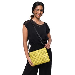 May include: A yellow crossbody bag with a black strap and a repeating pattern of black line art. The bag is held by a person wearing a black outfit.