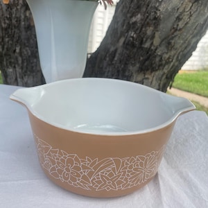 May include: A tan and white Pyrex bowl with a floral design. The bowl has a tan exterior with a white floral pattern around the base and a white interior. The bowl has two handles and is sitting on a white surface.