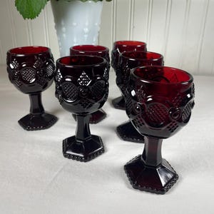 May include: A collection of six ruby red goblets with intricate, textured patterns. Each goblet features a hexagonal base and a detailed stem. A white vase with flowers is visible in the background.