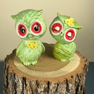 May include: Two ceramic owl figurines in a light green glaze. Each owl features large, prominent red-rimmed eyes with black pupils and small yellow flower accents on their chests or heads. The owls have textured, feather-like details.