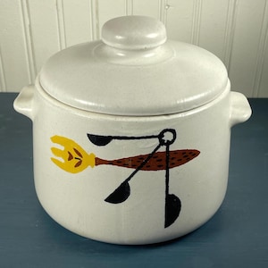 Peut inclure: Une cocotte en céramique de couleur crème avec un couvercle assorti et deux poignées latérales. L'extérieur présente un motif peint composé d'une fourchette jaune, d'un ustensile brun tacheté et de trois formes circulaires noires.