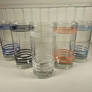 Puede incluir: Juego de cinco vasos altos y transparentes. Cada vaso presenta bandas horizontales en azul, negro, blanco y naranja. Los vasos están colocados sobre una superficie blanca sobre un fondo blanco.