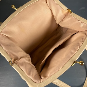 May include: Beige handbag with a structured frame and a kiss-lock clasp. The interior features a light pink lining with multiple compartments. The bag has a top handle and gold-toned hardware. The overall design is classic and elegant.