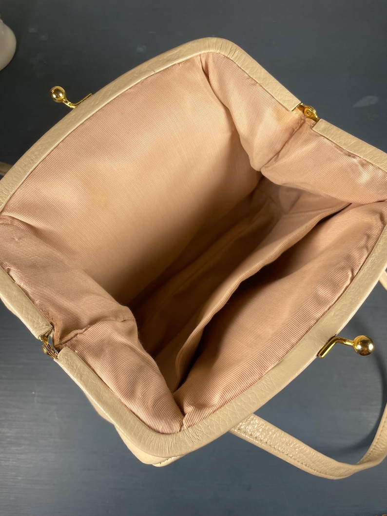 May include: Beige handbag with a structured frame and a kiss-lock clasp. The interior features a light pink lining with multiple compartments. The bag has a top handle and gold-toned hardware. The overall design is classic and elegant.