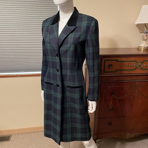 May include: A knee-length coat featuring a dark green and navy blue plaid pattern. The coat has a solid black velvet collar and matching black velvet trim on the two front pockets. It is fastened with three dark buttons down the center front.