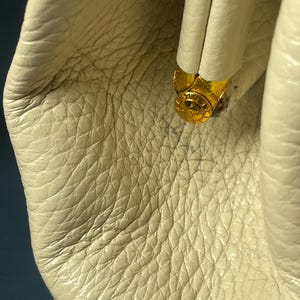 May include: Close-up of a cream-colored leather bag with a textured surface. The bag features a gold-colored metal clasp. The leather has a natural grain and subtle variations in color.
