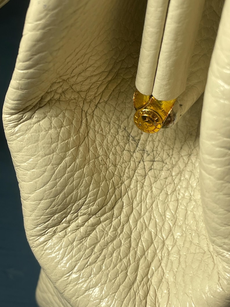 May include: Close-up of a cream-colored leather bag with a textured surface. The bag features a gold-colored metal clasp. The leather has a natural grain and subtle variations in color.