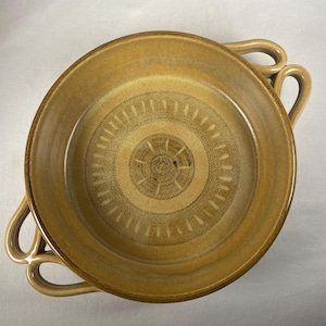 May include: A round, ceramic serving bowl with a tan glaze. The bowl features two loop handles and a concentric circle design in the center. The bowl is approximately 10 inches in diameter.