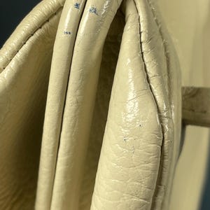 May include: Close-up of a cream-colored handbag with a textured leather exterior. The bag features a folded design with visible seams and a gold-colored clasp. The leather shows signs of wear and tear.