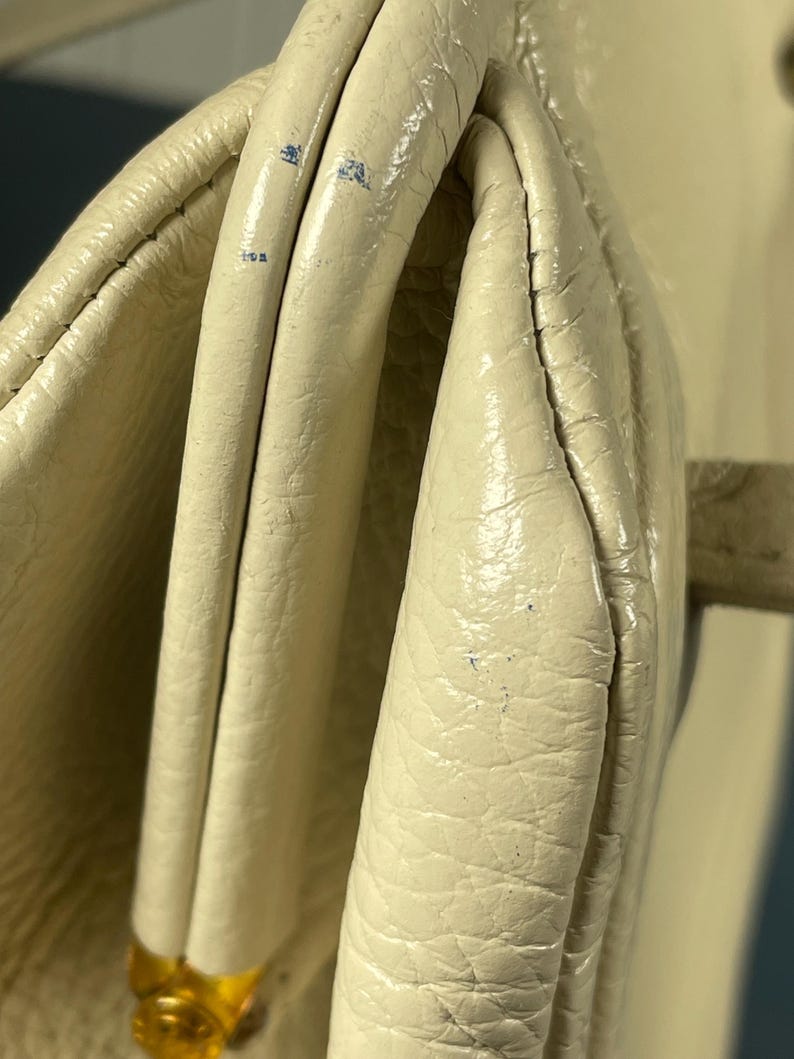 May include: Close-up of a cream-colored handbag with a textured leather exterior. The bag features a folded design with visible seams and a gold-colored clasp. The leather shows signs of wear and tear.