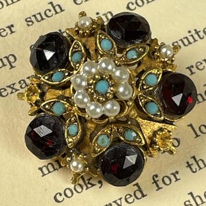 Könnte beinhalten: Vintage goldfarbene Brosche mit floralem Design. Die Brosche hat große, facettierte, dunkelrote und schwarze Edelsteine, kleine Türkissteine und weiße Perlen. Die Brosche ist vor dem Hintergrund von gealtertem Papier platziert.
