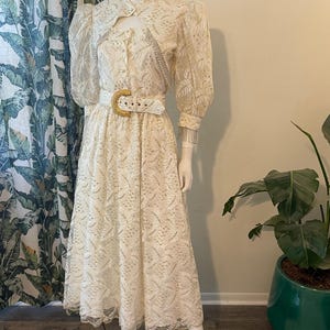 Op de afbeelding: Crèmekleurige vintage jurk met bijpassende bolero. De jurk heeft een bloemen kanten patroon, lange mouwen en een midi-rok. Een witte riem met een gouden gesp accentueert de taille. De bolero heeft een sleutelgat halslijn en zilveren ketting details.