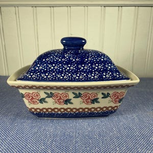 Pode incluir: Uma manteigueira de cerâmica com tampa. A tampa é azul escuro com bolinhas brancas e um pequeno botão. A base do prato é branca com um padrão floral de rosas vermelhas e folhas verdes, e uma borda marrom. O prato está sobre uma superfície azul estampada.