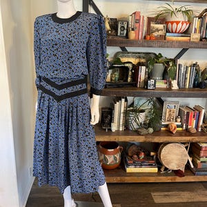 May include: A vintage blue dress with a paisley pattern and black trim. The dress features a round neckline, long sleeves, and a pleated skirt. The dress is displayed on a mannequin.