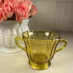 May include: A yellow glass sugar bowl with two handles and an embossed floral design. The bowl sits on a small pedestal base. A white vase with pink roses is in the background.
