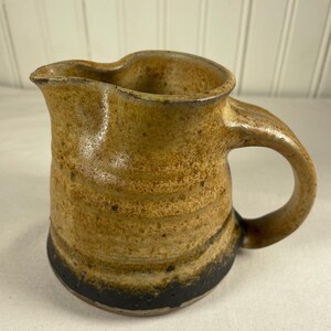May include: A handmade ceramic pitcher with a handle and spout. The pitcher has a textured, speckled surface with shades of brown and tan, and a black band at the base. The design is rustic and earthy.