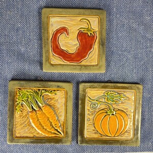 Puede incluir: Tres azulejos cuadrados de cerámica con un borde de bronce metálico. El azulejo superior presenta un chile rojo. El azulejo inferior izquierdo muestra tres zanahorias naranjas con hojas verdes. El azulejo inferior derecho muestra una calabaza naranja.