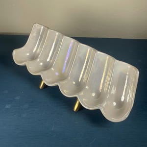 May include: A rectangular ceramic serving dish with a scalloped, iridescent white finish. The dish features five distinct rounded compartments and is supported by three small, cylindrical gold-toned metal legs.