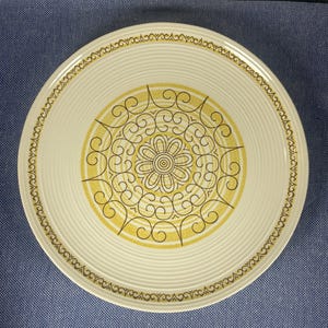 May include: Off-white ceramic plate with a yellow and brown floral design in the center. The plate has a repeating swirl pattern around the edge and a decorative border. The plate is on a blue background.