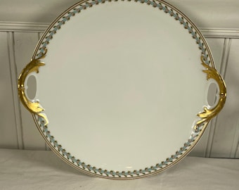 May include: A white porcelain serving platter with a gold rim and handles. The rim features a delicate blue and gold leaf pattern. The handles are ornate gold-colored, adding a touch of elegance to the serving dish.
