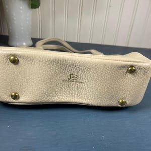 May include: A cream-colored leather handbag with a textured surface. The bag has a small handle and gold-colored metal feet. The brand name is visible on the front of the bag.