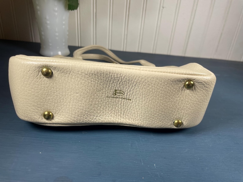 May include: A cream-colored leather handbag with a textured surface. The bag has a small handle and gold-colored metal feet. The brand name is visible on the front of the bag.