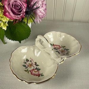 Könnte beinhalten: Cremefarbene, zweiteilige Servierschale mit Griff. Die Schale hat ein Blumendesign mit rosa und weißen Blumen und grünen Blättern. Die Ränder sind mit Gold verziert. Ein dekoratives Stück zum Servieren von Snacks oder kleinen Gegenständen.