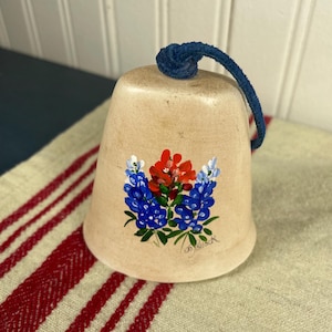 May include: A beige bell with a blue rope handle. The bell features a hand-painted floral design in red, blue, and white. The bell is sitting on a red and white striped cloth.