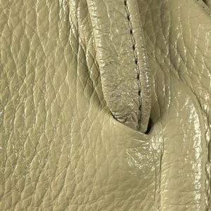 May include: Close-up of a beige leather handbag. The leather has a textured, pebbled appearance with visible stitching along the handle. The bag's surface reflects light, highlighting its glossy finish.
