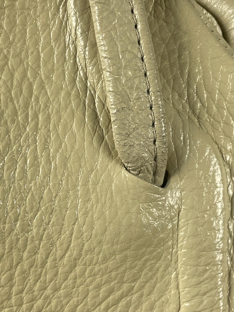 May include: Close-up of a beige leather handbag. The leather has a textured, pebbled appearance with visible stitching along the handle. The bag's surface reflects light, highlighting its glossy finish.