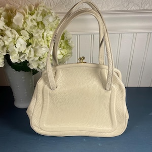 May include: Cream-colored vintage handbag with a structured design. The bag features a top handle, a gold-tone clasp, and a textured leather exterior. The bag is sitting on a blue surface.