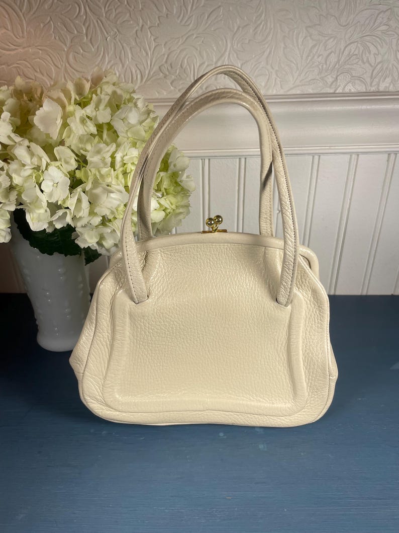 May include: Cream-colored vintage handbag with a structured design. The bag features a top handle, a gold-tone clasp, and a textured leather exterior. The bag is sitting on a blue surface.