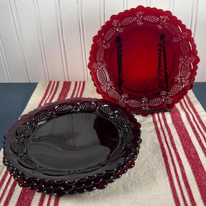 May include: A stack of four dark red glass plates with a scalloped edge and embossed floral design. A larger red glass plate with a similar design is displayed on a black metal stand. The plates are on a red and white striped cloth.