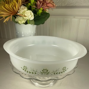 Może przedstawiać: Białe, okrągłe naczynie do zapiekania z zielonym kwiatowym wzorem, spoczywające na przezroczystym szklanym stojaku na ciasto. Naczynie ma prosty, elegancki design. W tle wazon z kwiatami.