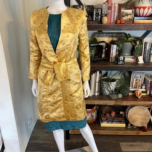 May include: A gold-colored, long-sleeved coat with a floral pattern and a matching belt is displayed on a mannequin. The coat is paired with a teal-colored dress. The mannequin is positioned in front of a wooden bookshelf filled with books and decorative items.