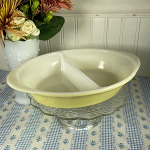 May include: A vintage, oval-shaped divided serving dish in a pale yellow color. The dish has a white divider and is displayed on a clear glass cake stand. The background includes a vase of flowers and a blue and white checkered tablecloth.