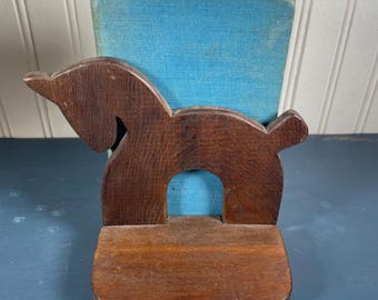 May include: A dark brown wooden bookend in the shape of a horse. The bookend features a curved base and a central cutout. A stack of books with blue covers is held in the bookend.