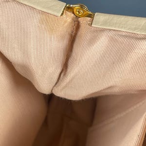 May include: Close-up of a vintage handbag interior. The bag features a light beige exterior trim and a gold-colored clasp. The interior is a light peach color, with a textured fabric lining. The bag appears to be made of a soft material.