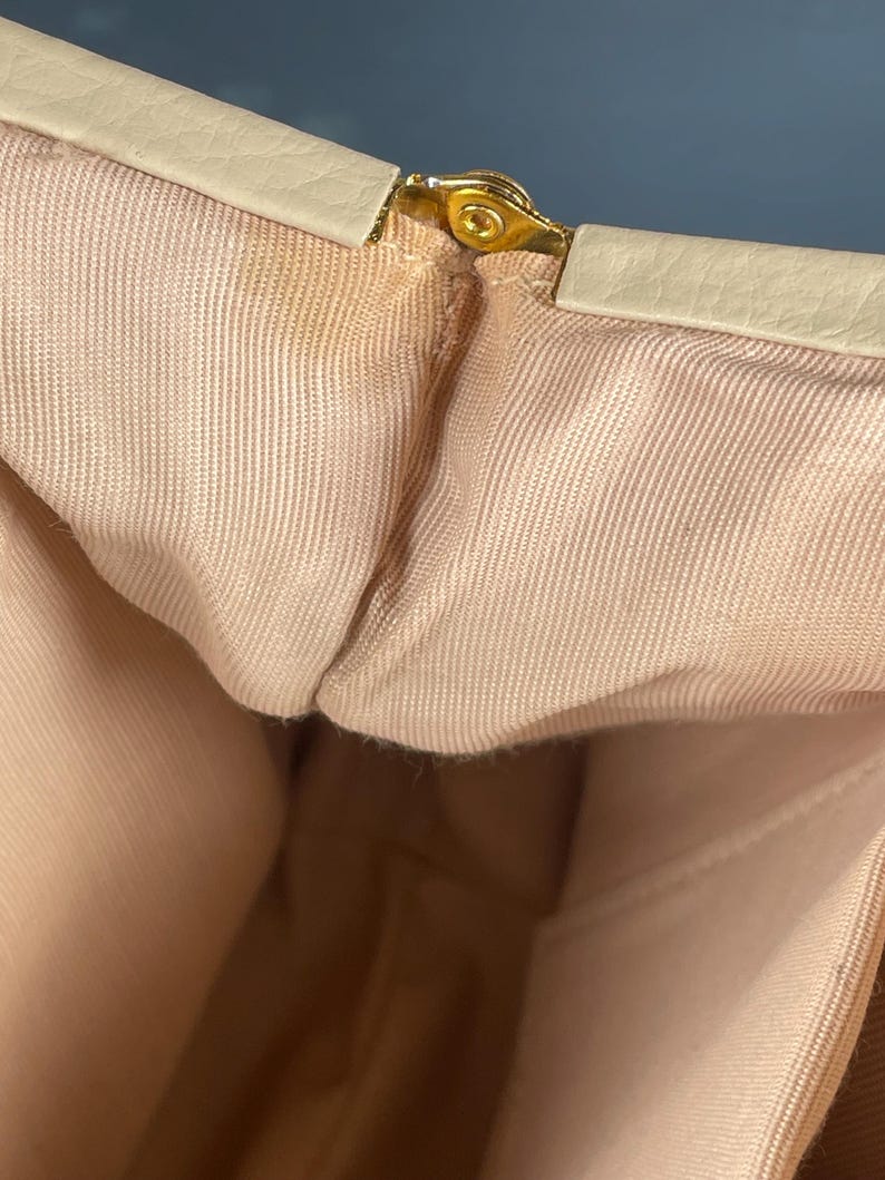 May include: Close-up of a vintage handbag interior. The bag features a light beige exterior trim and a gold-colored clasp. The interior is a light peach color, with a textured fabric lining. The bag appears to be made of a soft material.