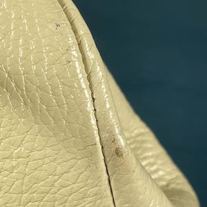 May include: Close-up of a section of a cream-colored leather bag. The leather has a textured, pebbled surface with visible stitching along the seam. The bag appears to be slightly worn, with minor discoloration.