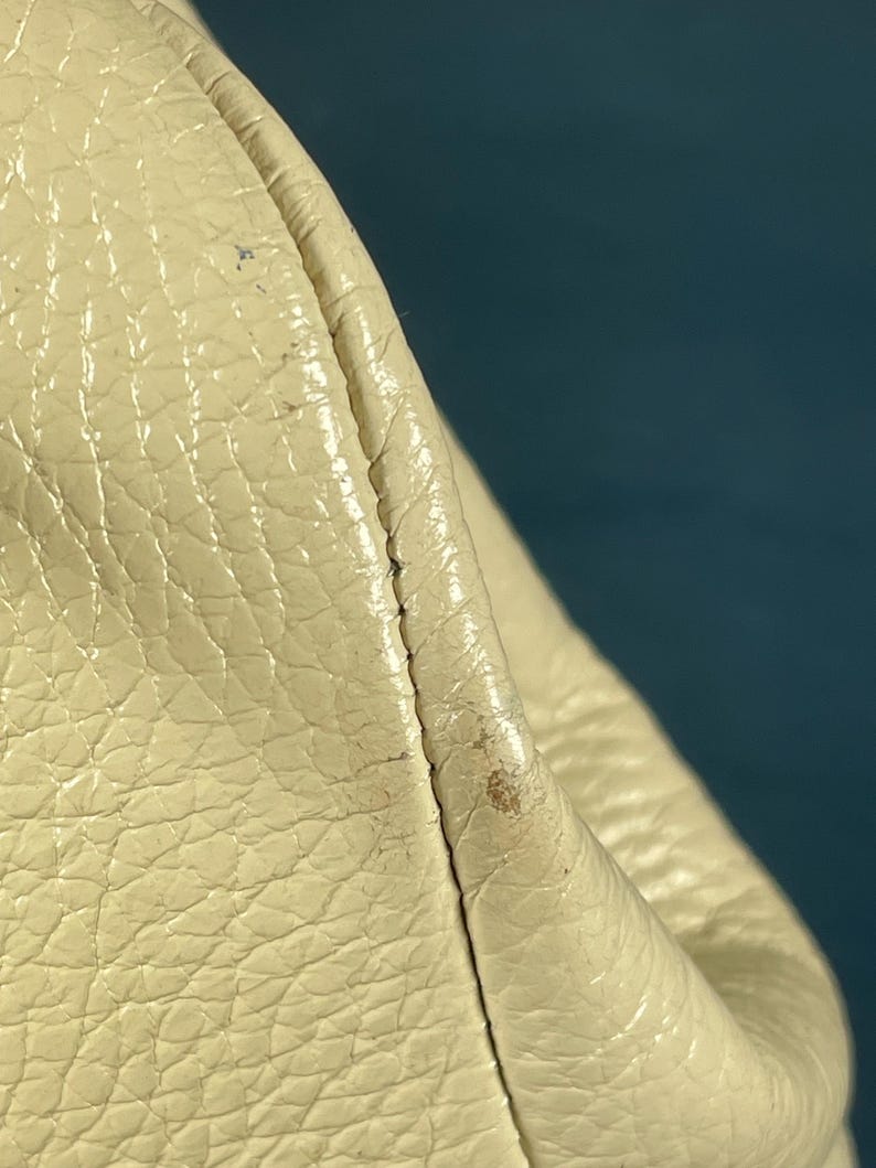 May include: Close-up of a section of a cream-colored leather bag. The leather has a textured, pebbled surface with visible stitching along the seam. The bag appears to be slightly worn, with minor discoloration.