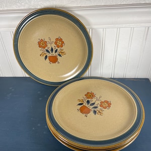 Puede incluir: Juego de platos de cerámica con centro beige, borde azul marino y borde exterior marrón. Cada plato presenta un diseño floral con flores naranjas y un jarrón marrón. Los platos están apilados sobre una superficie azul oscuro.