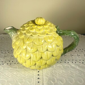 May include: A ceramic teapot shaped like a yellow artichoke with a matching lid. The body features overlapping yellow petal-like scales, while the handle and spout are textured and colored in a deep green to resemble the stem and leaves of the plant.
