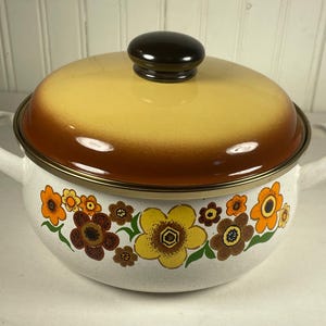 May include: A vintage white enamel pot with a lid. The pot features a floral design in orange, yellow, and brown. The lid is yellow and brown with a dark brown knob. It has two white handles and a gold rim.