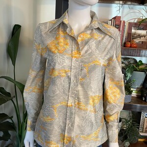 Op de afbeelding: Een button-down shirt met lange mouwen met een grijs en geel bloemenpatroon. Het shirt wordt op een mannequin gepresenteerd. Het heeft een kraag en manchetten met knopen. De achtergrond omvat planten en een houten plank.
