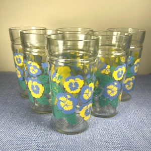 May include: A set of six clear glass tumblers featuring a repeating pattern of blue and yellow pansy flowers with green leaves. Each glass has a cylindrical shape with a slightly wider rim and a smooth, transparent finish throughout the body.