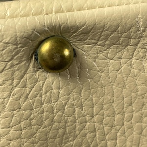 May include: Close-up of a cream-colored leather surface with a brass-colored stud. The leather has a textured, pebbled appearance. The stud is round and slightly domed, with a visible indentation where it is attached.