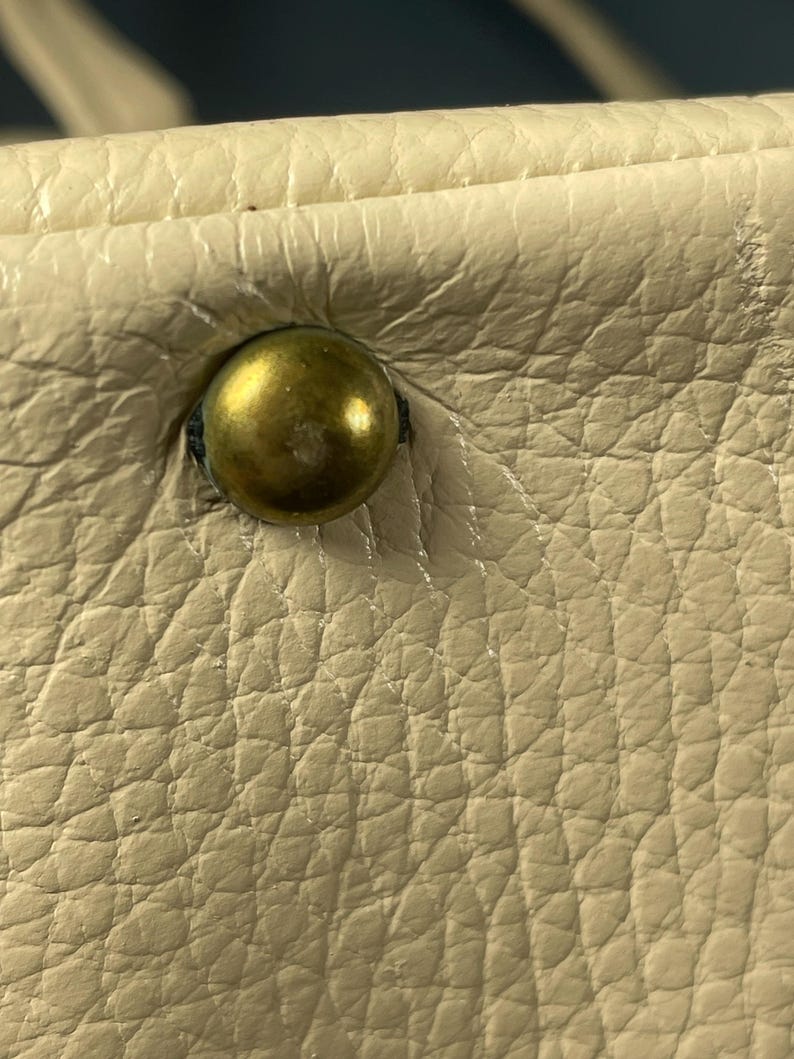 May include: Close-up of a cream-colored leather surface with a brass-colored stud. The leather has a textured, pebbled appearance. The stud is round and slightly domed, with a visible indentation where it is attached.