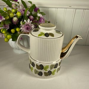 May include: Cream-colored vintage teapot with a gold-colored spout and handle. The teapot has a ribbed design on the upper portion and a leaf pattern on the lower portion. The lid also features a leaf pattern. A bouquet of flowers sits to the left of the teapot.